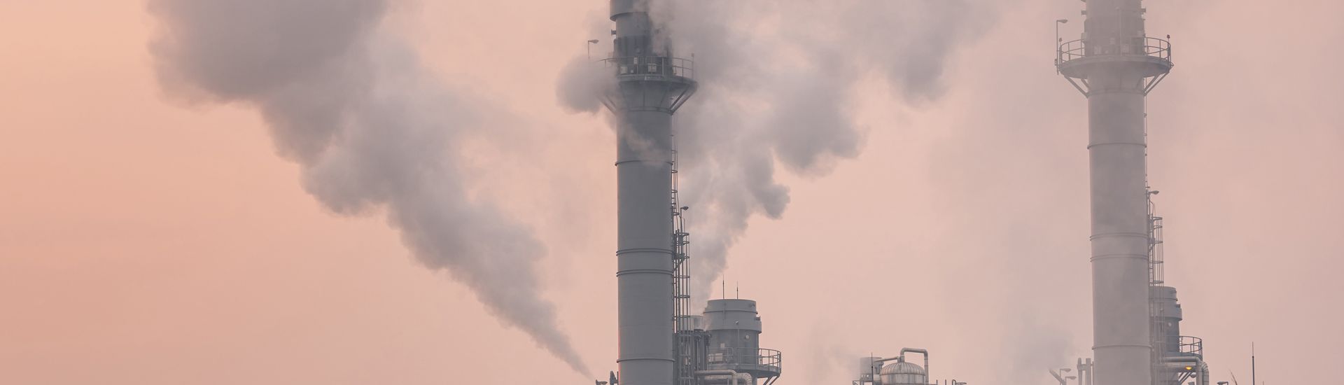 Factory with plumes of smoke coming out of the top, against a hazy pink sky.